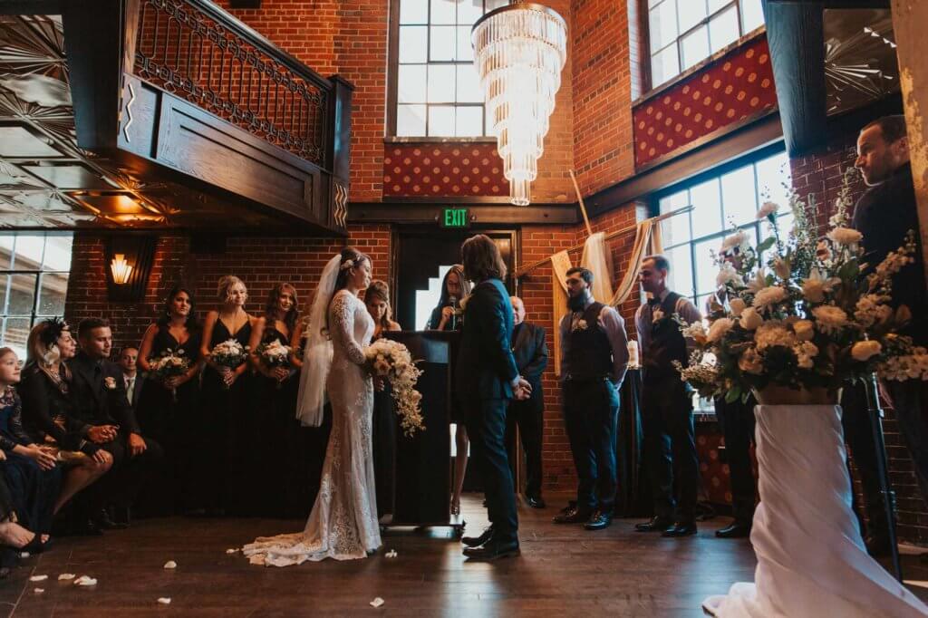 Bride and groom standing at the altar surrounded by wedding guests and floral arrangements during an indoor ceremony at Ironworks Denver.